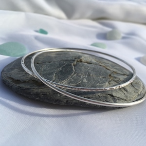 May include: Two silver bangles, one with a hammered texture, stacked on a grey stone.