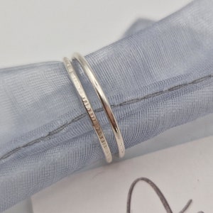 May include: Two thin silver rings stacked on top of each other. The top ring has a textured finish. The rings are on a light blue fabric background. The text "Anellas Jewellery Box" is visible in the background.