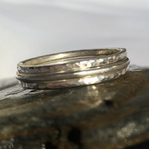 May include: Three silver stacking rings with a hammered texture. The rings are stacked on a grey rock.