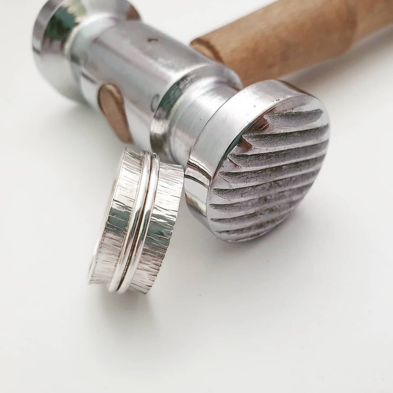 Pode incluir: Um anel de prata com um acabamento martelado texturizado. O anel &eacute; feito de duas bandas que est&atilde;o entrela&ccedil;adas. O anel &eacute; mostrado ao lado de um martelo de metal com uma cabe&ccedil;a texturizada.