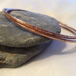 May include: Two hammered copper bangles stacked on top of each other. The bangles are a warm rose gold colour.
