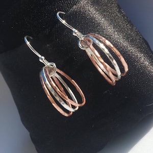 Pode incluir: Um par de brincos pendurados com três argolas texturizadas em tons de prata e ouro rosa. Os brincos têm um fecho de gancho simples e são exibidos em uma superfície preta.