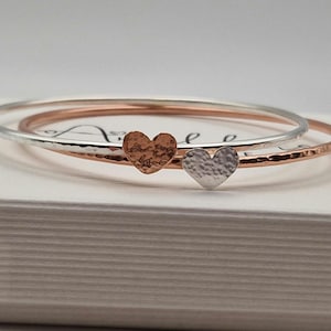 Pode incluir: Dois braceletes de metal martelado, um prateado e outro de ouro rosa, com pingentes em forma de coração. Os pingentes são texturizados e têm um acabamento martelado.