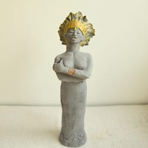 Könnte beinhalten: Eine graue Skulptur einer stehenden Figur mit vor der Brust verschränkten Armen. Die Figur trägt einen strukturierten, goldgelben Kopfschmuck und ein goldfarbenes Armband am Handgelenk. Der Gesichtsausdruck ist neutral mit geschlossenen Augen.