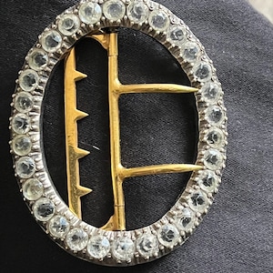 May include: Oval-shaped antique buckle with a silver-colored frame and a gold-colored clasp. The frame is encrusted with clear, round gemstones. The buckle's design is symmetrical, with a central gold bar and a series of teeth.