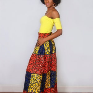 May include: A woman wearing a yellow off-the-shoulder top and a long, colourful skirt with a geometric pattern. The skirt is made of a patchwork of red, blue, and yellow fabric with a gold swirl design.