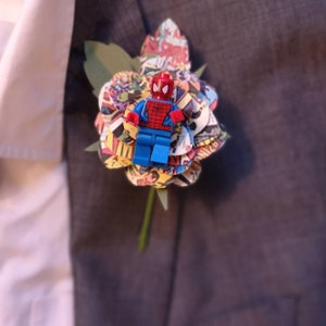 Op de afbeelding: Een unieke corsage met een Lego Spider-Man figuur bovenop een bloem gemaakt van stripboekpagina's. De bloem heeft groene bladeren en een groene stengel, bevestigd aan een grijs pak.