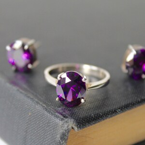 May include: A set of jewelry featuring a silver ring with a large oval-shaped purple gemstone and two matching stud earrings with smaller purple gemstones.