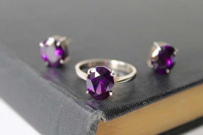 May include: A set of jewelry featuring a silver ring with a large oval-shaped purple gemstone and two matching stud earrings with smaller purple gemstones.