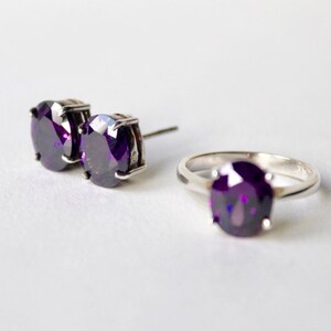 May include: A set of purple gemstone jewelry featuring a ring and a pair of earrings. The ring and earrings have a silver setting.