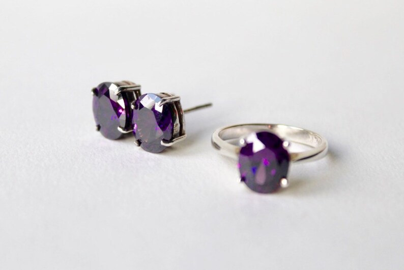 May include: A set of purple gemstone jewelry featuring a ring and a pair of earrings. The ring and earrings have a silver setting.