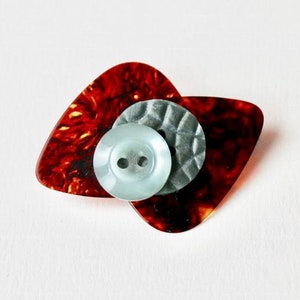 以下が含まれることがあります： 上面が2色のボタンが付いたギターピック。ピックは茶色で、べっ甲模様。ボタンは水色で、穴が2つあり、周りを銀色の輪っかが囲んでいる