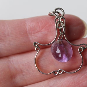 May include: A silver charm with a delicate, openwork design in the shape of a hand holding a round, purple amethyst stone.