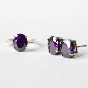 May include: A set of jewelry featuring a ring and a pair of earrings. Both the ring and earrings have a large, oval-shaped, dark purple gemstone.