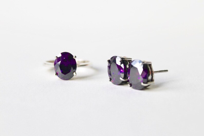 May include: A set of jewelry featuring a ring and a pair of earrings. Both the ring and earrings have a large, oval-shaped, dark purple gemstone.