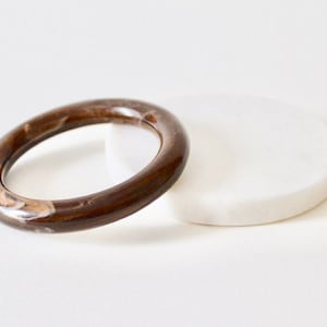 May include: A brown and tan marbled bangle bracelet. The circular bracelet is made of a glossy material and rests on a white, circular surface. The bangle is a stylish accessory.