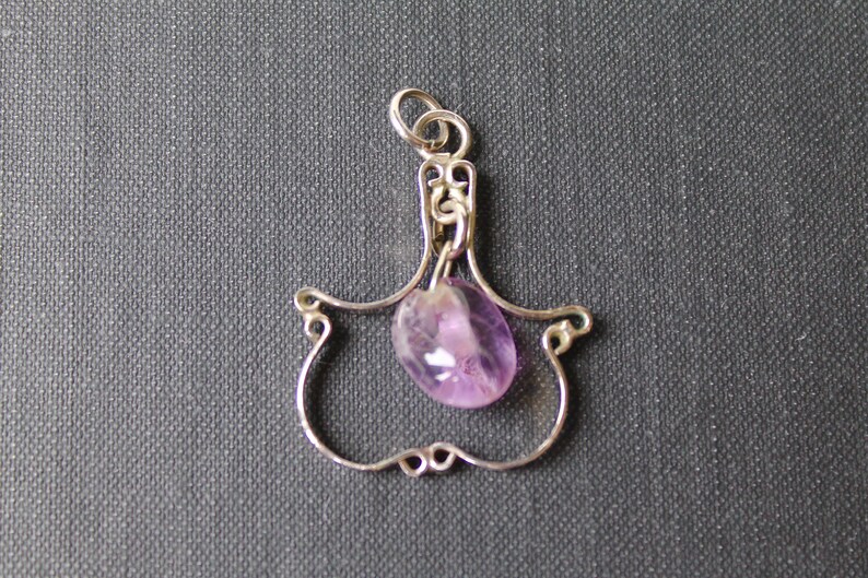 May include: A silver pendant with a delicate, ornate design features a dangling amethyst gemstone. The pendant is shaped like a teardrop and has a loop at the top for attaching to a chain.