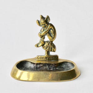 May include: A brass trinket dish featuring a small, intricate figure of a pixie or gnome sitting on top. The dish is oval shaped and has the word "CORNWALL" inscribed on the front.