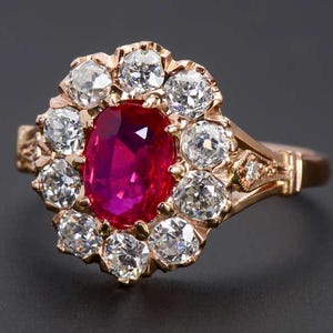 May include: A rose gold ring featuring a central oval-cut vibrant red gemstone surrounded by a halo of ten round-cut clear white stones. The metal band displays intricate engraved detailing along the shoulders, leading to a polished solid shank.