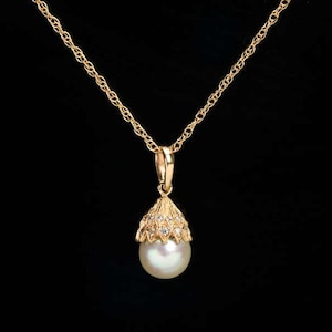 May include: A gold-colored necklace with a pendant featuring a white pearl. The pendant is encased in a decorative gold setting adorned with small, clear gemstones. The chain is a delicate, twisted design, set against a black background.