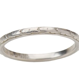 May include: A thin silver-toned metal ring featuring a continuous engraved pattern of small, stylized floral or cloud-like shapes around the entire outer circumference of the band. The metal has a smooth, polished finish with visible etched details.