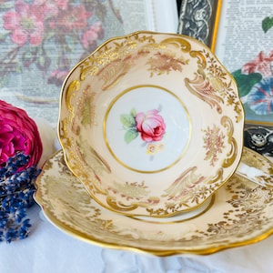 Könnte beinhalten: Ein Vintage-Teetassen- und Untertassen-Set mit einem zarten Blumendesign und goldenen Akzenten. Die Tasse zeigt eine rosa Rose mit grünen Blättern und einem weißen Hintergrund. Die Untertasse hat ein ähnliches Blumenmuster mit goldenen Verzierungen.