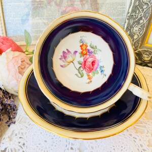 May include: A vintage teacup and saucer set. The teacup and saucer have a navy blue border with a gold trim. The center of the teacup features a floral design with pink roses, purple tulips, and other colorful flowers. The set is placed on a white lace tablecloth.