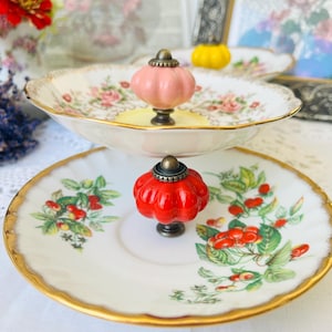 Op de afbeelding: Een serveerstandaard met twee niveaus met vintage borden met bloemenpatroon en gouden rand. De standaard is versierd met een roze en een rode decoratieve knop. De borden zijn versierd met bloemen- en bessenontwerpen.
