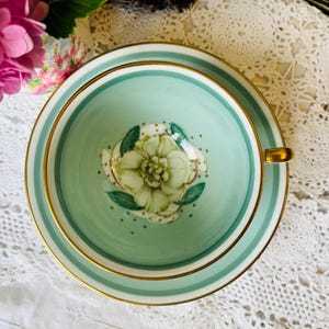 May include: A vintage teacup and saucer set. The cup and saucer are a light teal color with gold trim. The inside of the cup features a white floral design with green leaves and gold accents. The saucer has a matching design.