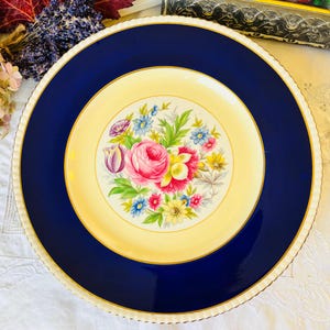 Puede incluir: Un plato grande, redondo, de color crema con un borde azul y un ribete dorado. El centro del plato presenta un diseño floral con flores rosas, amarillas y azules.