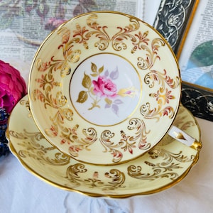 Puede incluir: Un juego de taza y platillo de té vintage con un delicado diseño floral. La taza y el platillo son amarillos con detalles dorados y una rosa rosa en el centro.
