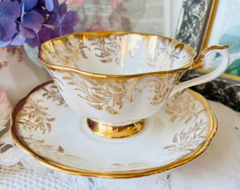 May include: A white porcelain teacup and saucer set featuring a delicate gold leaf pattern. The teacup has a scalloped rim, a gold-trimmed base, and a curved handle. The saucer matches with a scalloped edge and gold botanical motifs throughout.