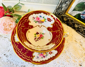 May include: A vintage teacup and saucer set. The set features a burgundy and gold design with floral accents. The teacup and saucer have a scalloped edge. The teacup has a small handle. The set is on a white lace tablecloth.