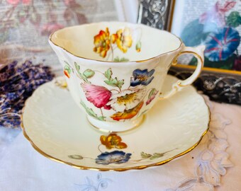 Puede incluir: Juego de taza y platillo de color crema con un diseño floral con flores rojas, amarillas y azules. La taza tiene un borde festoneado y un asa dorada. El platillo tiene un patrón floral a juego y un borde dorado. Un juego de té vintage.