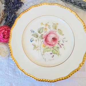 May include: A white porcelain dinner plate with a gold rim and a floral design featuring a pink rose and other flowers. The plate has the word "Crown" printed on the bottom.