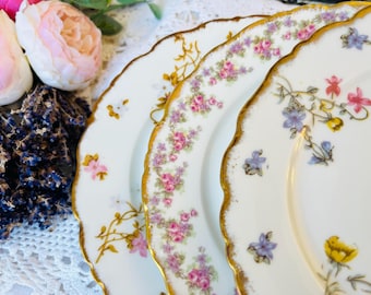 May include: Close-up of three white porcelain plates with scalloped edges and gold trim. The plates are decorated with floral patterns in pink, purple, and yellow. The plates are arranged in a slight arc, showcasing their intricate designs.