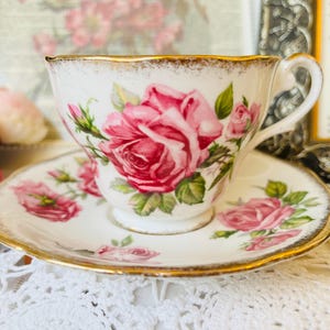 May include: A white teacup and saucer set with a gold rim and a floral rose design. The teacup and saucer feature a large pink rose and smaller pink rose buds with green leaves. The set is on a white lace surface.