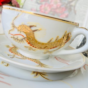 May include: White porcelain teacup and saucer set with gold dragon and trim details. The dragon design is in gold and red. The cup has a curved handle. The saucer is oval-shaped.