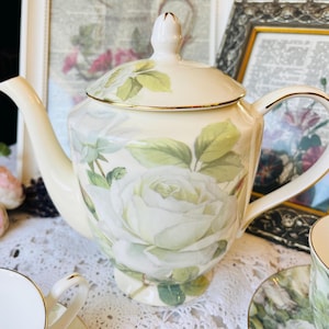 May include: Cream porcelain teapot featuring a large white rose and green leaf design. The teapot has a gold trim and a matching lid. The handle and spout are curved. A matching teacup and saucer are visible.