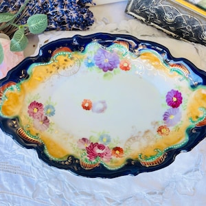 Op de afbeelding: Ovale antieke serveerschaal met een geschulpte rand. De schaal heeft een wit interieur met bloemmotieven in roze, paars en blauw. De rand is marineblauw, met gouden en gele accenten. Een decoratief stuk voor het serveren van voedsel.
