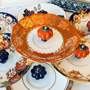 Peut inclure: Une collection d'assiettes décoratives vintage avec des accents floraux et dorés. Les assiettes présentent une variété de couleurs, dont l'orange, le bleu et le blanc. Chaque assiette a un bouton décoratif au centre. Les assiettes sont disposées sur une surface blanche.