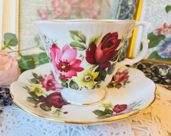 May include: A white teacup and saucer set with a floral design. The cup and saucer are decorated with red and pink roses, yellow flowers, and green leaves. The rims are trimmed with gold. A classic tea set.