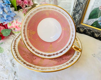 May include: A porcelain teacup and saucer set featuring a soft pink interior and rim, decorated with intricate gold filigree patterns. The teacup has a white center and a gold-trimmed handle, while the saucer matches the pink and gold design.