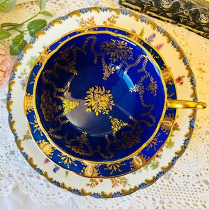 Peut inclure: Service à thé vintage composé d'une tasse et d'une soucoupe. La tasse est bleu foncé avec des motifs floraux dorés et une anse dorée. La soucoupe est blanche avec des bordures florales et dorées.