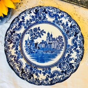 May include: A blue and white ceramic plate with a decorative, scalloped edge. The plate features a central landscape scene with buildings and water, surrounded by floral patterns. A classic design.