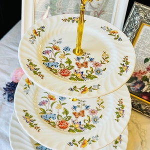 May include: A three-tiered serving stand with a gold handle. Each white plate features a floral design with red, blue, yellow, and pink flowers, and butterflies. The plates have a swirled edge.