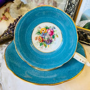 May include: A teal blue teacup and saucer set with a floral design in the centre of the saucer. The teacup has a gold rim and the saucer has a gold rim and a gold band around the edge.