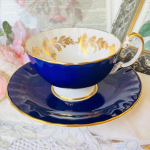 Peut inclure: Un ensemble tasse et soucoupe en porcelaine avec un extérieur bleu cobalt profond, des bords festonnés et une bordure dorée. L'intérieur de la tasse est blanc avec un motif de feuilles dorées sur le bord, et l'anse est blanche avec des détails dorés.