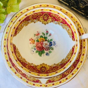 Puede incluir: Un juego de taza y platillo de té vintage con un diseño floral. La taza y el platillo son blancos con un borde rojo y dorado. El diseño floral presenta un ramo de flores rosas, amarillas y blancas.