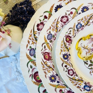 May include: Set of three white ceramic plates with a floral design featuring red, purple, and yellow flowers. The plates have a gold rim and a gold handle on the largest plate.